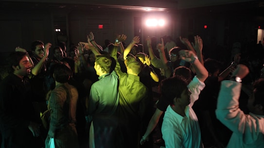 A group of people is energetically dancing in a dimly lit room. The atmosphere is lively, with individuals raising their hands and moving to the music. Soft green lighting and a few bright spotlights create a vivid contrast.