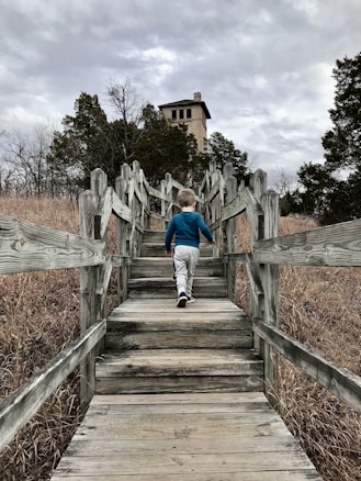 A child is walking up a wooden pathway surrounded by dry grass and trees, leading towards a stone building that resembles a tower. The sky is overcast with dense, gray clouds.