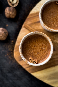 Two cups filled with a brown liquid, possibly hot chocolate or coffee, are placed on a wooden surface. The liquid is topped with a powdery substance, likely cinnamon. There are also some whole walnuts or nutmeg scattered around.