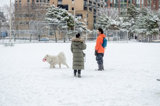 man in brown coat standing on snow covered ground with white dog during daytime