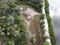 An aerial view of a wooded area featuring a ropes course with platforms and poles. The ropes course is located near a paved path that runs alongside a body of water. Lush greenery and trees surround the area, creating a natural and serene environment.