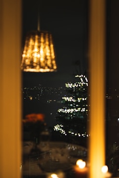 A warm, dimly lit interior with a hanging lamp softly glowing. Outside the window, a cityscape at night is visible with a Christmas tree adorned with twinkling lights. Candles and blurred floral arrangements add to the cozy ambience.