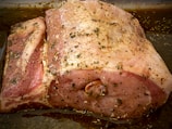 A large piece of raw pork, seasoned with herbs and spices, lies in a baking tray. The meat is marbled, showcasing the fat layers and is covered with a mix of green herbs.