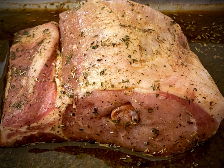 A large piece of raw pork, seasoned with herbs and spices, lies in a baking tray. The meat is marbled, showcasing the fat layers and is covered with a mix of green herbs.