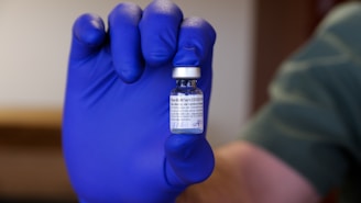 A gloved hand holds a small vial labeled as a COVID-19 vaccine. The focus is on the vial, which is being securely grasped, with text visible on the label. The glove is a prominent bright blue color, contrasting with the neutral background.