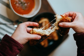 Two hands holding a piece of grilled cheese sandwich with melted cheese stretching between them. A bowl of tomato soup with herbs is visible in the background on a wooden table, alongside several toasted bread pieces.