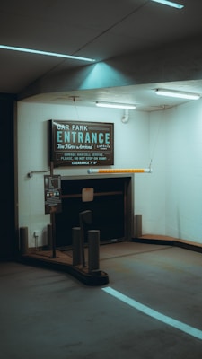 A dimly lit underground car park entrance with a prominent signboard displaying details about the parking garage, including clearance height and service information. The environment features white walls, ceiling lights, and a partially visible payment machine.