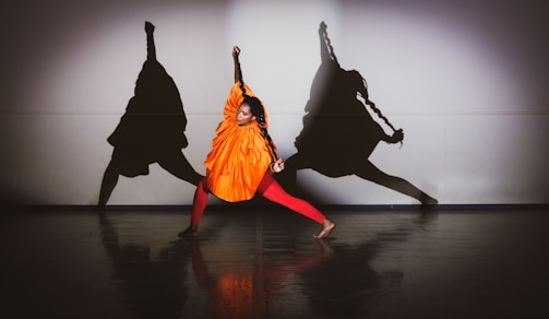 A person dressed in a bright orange outfit and red leggings strikes a dynamic pose in a dance studio. Three large shadows are cast dramatically on the wall behind, adding a sense of movement and depth.