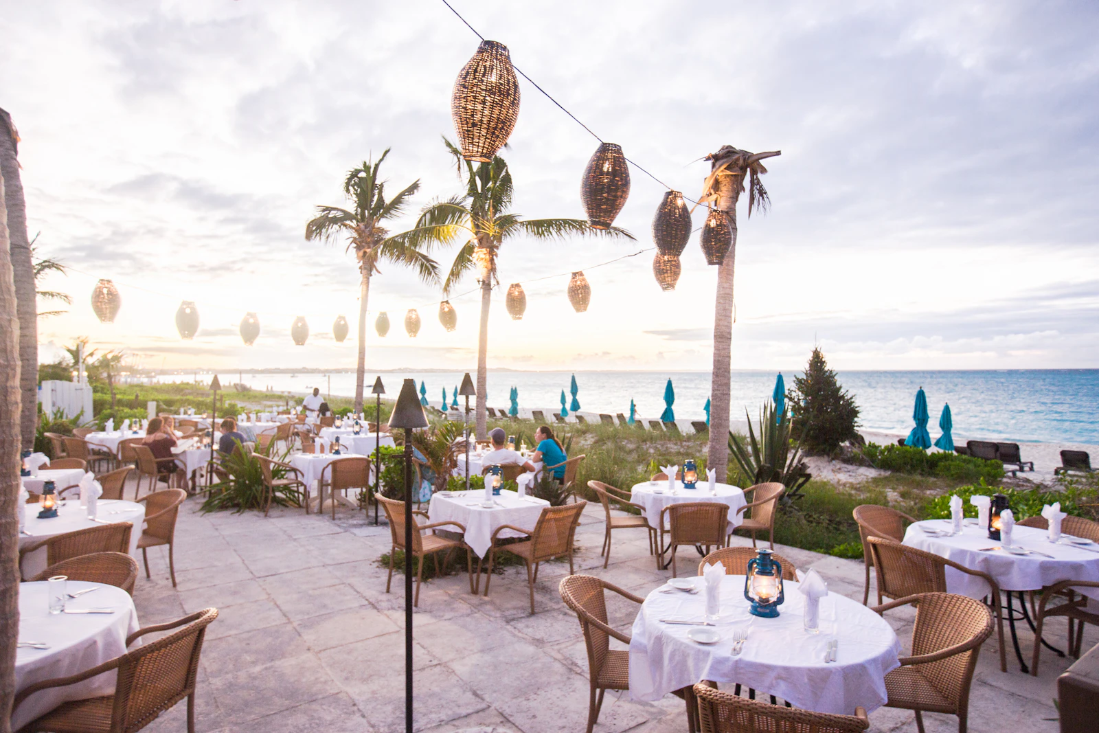 An oceanfront terrace set with white-linen tables and hanging lanterns beside palm trees