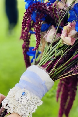 A vibrant bouquet featuring blue and pink flowers tied with a purple ribbon. There is a lace detail and a charm with a date and names written on it.