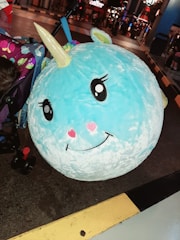 A large, plush toy shaped like a smiling, blue unicorn with a golden horn and pink heart-shaped cheeks is propped against a stroller adorned with colorful butterfly patterns. The background reveals an outdoor or amusement park setting with people walking and lights glowing in the distance.