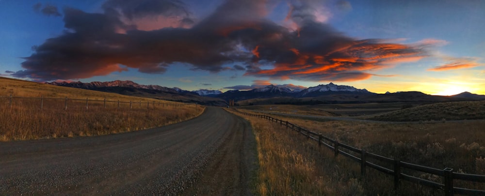 gray asphalt road between green grass field under orange and gray cloudy sky