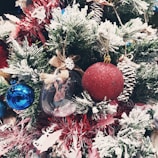 A festive Christmas tree adorned with a variety of decorations, including a glittery red bauble, a shiny blue ornament, pine cones dusted with fake snow, and a small figurine encased in a clear globe. The branches are lush and green, with a layer of artificial snow, adding a wintery touch.
