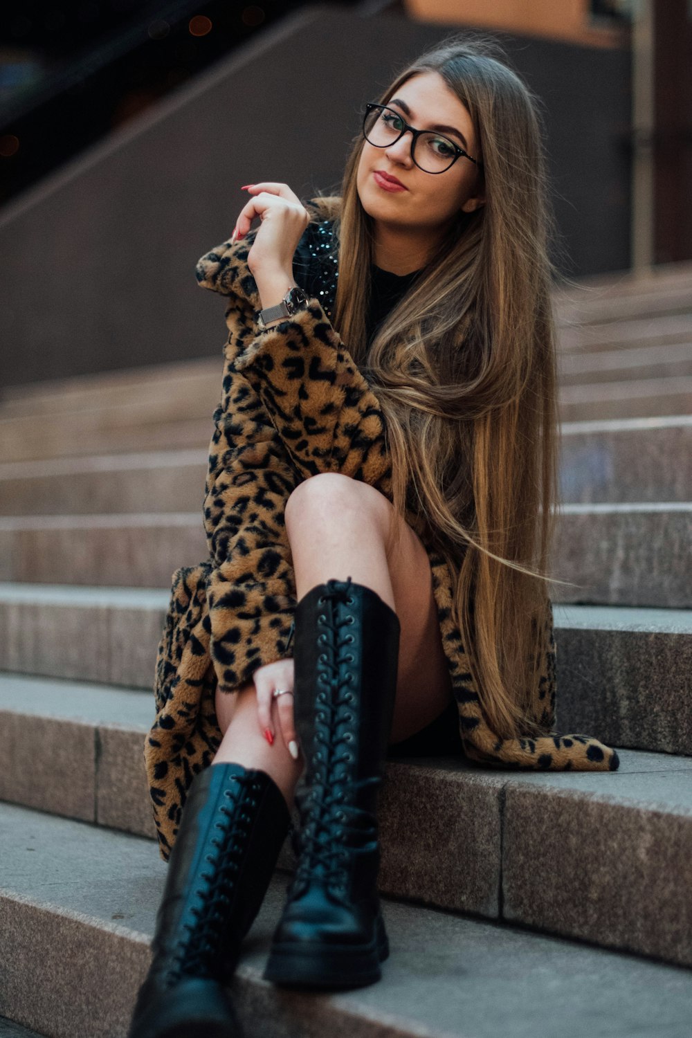 Mujer con abrigo con estampado de leopardo y botas negras sentada en escaleras de hormigón