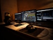 Three laptop screens are open on a desk, showing different software applications related to music and video production. A pair of headphones, several small electronic devices, and a computer mouse are visible on the desk. The lighting is dim, creating a focused and concentrated atmosphere.