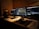 Three laptop screens are open on a desk, showing different software applications related to music and video production. A pair of headphones, several small electronic devices, and a computer mouse are visible on the desk. The lighting is dim, creating a focused and concentrated atmosphere.