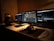 Three laptop screens are open on a desk, showing different software applications related to music and video production. A pair of headphones, several small electronic devices, and a computer mouse are visible on the desk. The lighting is dim, creating a focused and concentrated atmosphere.
