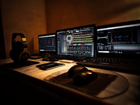 Three laptop screens are open on a desk, showing different software applications related to music and video production. A pair of headphones, several small electronic devices, and a computer mouse are visible on the desk. The lighting is dim, creating a focused and concentrated atmosphere.