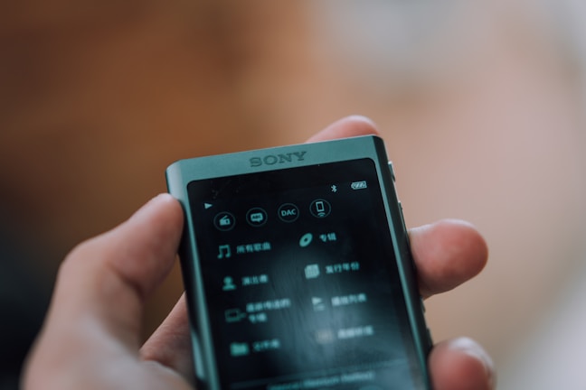 A person holds a small, portable Sony device, likely a music player, with a screen displaying an interface in a language that could be Chinese. The device has several options visible such as music, playlists, and more, indicating its function as a media player. The background is blurred and the focus is on the device.