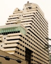 A tall, modern multi-story hotel building with a beige and brown facade. The structure has a unique design with staggered balconies and large windows near the top floor. The name 'Century Plaza Hotel' is visible on the upper section, accompanied by Chinese characters. Part of another building with a glass section is seen in the foreground.