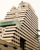 A tall, modern multi-story hotel building with a beige and brown facade. The structure has a unique design with staggered balconies and large windows near the top floor. The name 'Century Plaza Hotel' is visible on the upper section, accompanied by Chinese characters. Part of another building with a glass section is seen in the foreground.