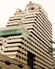 A tall, modern multi-story hotel building with a beige and brown facade. The structure has a unique design with staggered balconies and large windows near the top floor. The name 'Century Plaza Hotel' is visible on the upper section, accompanied by Chinese characters. Part of another building with a glass section is seen in the foreground.