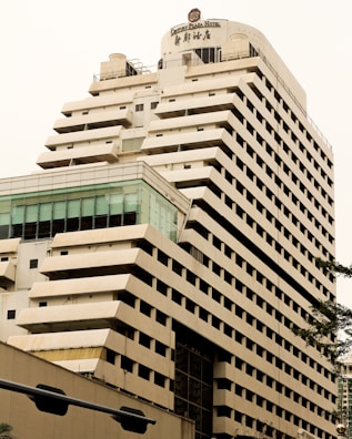 A tall, modern multi-story hotel building with a beige and brown facade. The structure has a unique design with staggered balconies and large windows near the top floor. The name 'Century Plaza Hotel' is visible on the upper section, accompanied by Chinese characters. Part of another building with a glass section is seen in the foreground.