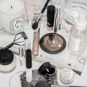 A collection of makeup and skincare products are displayed on a white surface. There are various items including a candle, makeup brushes, a bronze compact, a setting spray, and several cream and liquid cosmetics. Some products are from recognizable brands, and there is a book used as a base for the arrangement. An eyelash curler and rings are also part of the composition.