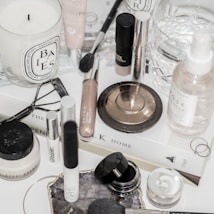 A collection of makeup and skincare products are displayed on a white surface. There are various items including a candle, makeup brushes, a bronze compact, a setting spray, and several cream and liquid cosmetics. Some products are from recognizable brands, and there is a book used as a base for the arrangement. An eyelash curler and rings are also part of the composition.