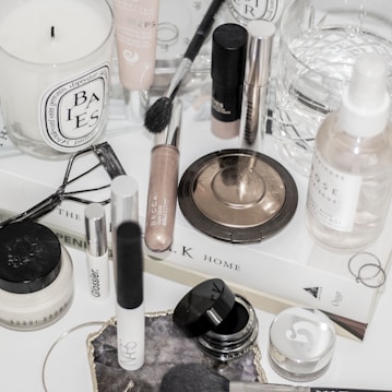 A collection of makeup and skincare products are displayed on a white surface. There are various items including a candle, makeup brushes, a bronze compact, a setting spray, and several cream and liquid cosmetics. Some products are from recognizable brands, and there is a book used as a base for the arrangement. An eyelash curler and rings are also part of the composition.