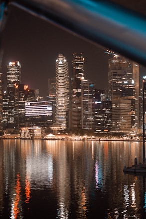 A vibrant cityscape at night with skyscrapers illuminated by various lights, reflecting off the calm water below. The scene captures modern architecture and the bustling yet tranquil atmosphere of an urban skyline.