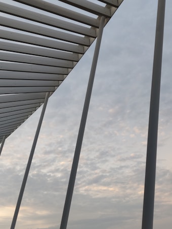 A series of large white beams form a modern architectural structure set against a sky filled with soft, light clouds. The perspective captures the beams converging toward a high point, creating a geometric pattern.