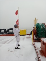 A person wearing a white jumpsuit and a hat is standing on a snowy deck of a ship, raising a red flag. The deck is covered with a thin layer of snow, and there are footprints visible. There is a large green-covered object to the right and a yellow metal box labeled 'ETA' in the background.