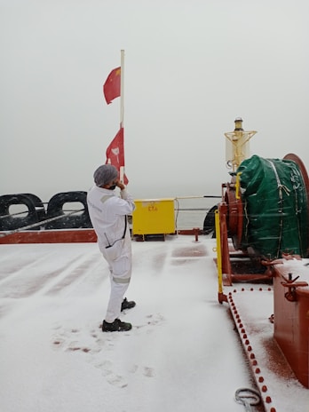 A person wearing a white jumpsuit and a hat is standing on a snowy deck of a ship, raising a red flag. The deck is covered with a thin layer of snow, and there are footprints visible. There is a large green-covered object to the right and a yellow metal box labeled 'ETA' in the background.