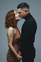 A man and a woman standing closely, holding hands, and about to kiss. The woman has curly red hair and is wearing a brown dress, while the man has short dark hair and is dressed in a black turtleneck.