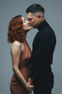 A man and a woman standing closely, holding hands, and about to kiss. The woman has curly red hair and is wearing a brown dress, while the man has short dark hair and is dressed in a black turtleneck.