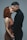 A man and a woman standing closely, holding hands, and about to kiss. The woman has curly red hair and is wearing a brown dress, while the man has short dark hair and is dressed in a black turtleneck.