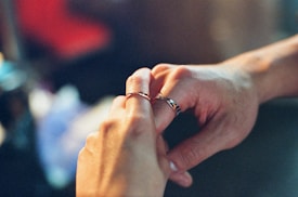 A close-up of two hands exchanging rings, with the focus on the hands and rings. The background is blurred, giving an intimate and personal feeling.