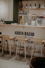 A cozy bar area featuring three wooden stools in front of a counter with a sign reading 'Green Bazar.' The counter has beer taps and is backed by shelves holding bottles, glasses, and a coffee grinder. The overall setting suggests a casual and inviting atmosphere.