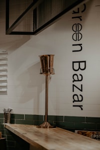 An indoor scene with a minimalist design featuring a tall, metallic stand placed on a light wooden table. The background has white tiled walls with black text reading 'Green Bazaar'. On the left, there is a metal container holding what appears to be straws or similar objects.