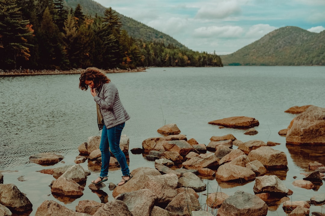 woman in gray jacket and blue denim jeans standing on brown rock near body of water