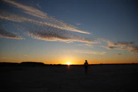 A sunset silhouette of a woman in a western jacket walking through open prairie.