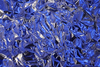 A crumpled sheet of metallic foil with a blue tint, displaying a complex texture of wrinkles and folds. The surface reflects light, creating a shiny effect with contrasting areas of shadow and brightness.