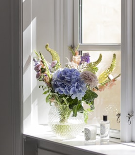 purple and white flowers in white ceramic vase beside window