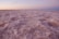 A vast expanse of pink salt crystals stretches out under a dusky sky. The surface is uneven and has a textured appearance, reminiscent of a frozen landscape. The horizon is barely visible in the distance, with soft hues of orange and pink coloring the sky.