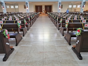 An indoor space with rows of wooden pews, decorated with red and green ornaments, and marked with red X signs for seating restrictions. The area appears to be well-lit with large doors at the far end and a few people present, including one person standing near the entrance.