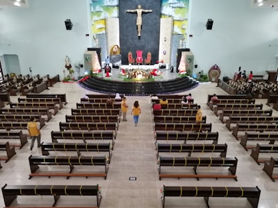 A large church interior featuring wooden benches with yellow ribbons, a central aisle, and a raised altar decorated with flowers and red cloth. A prominent statue of Jesus is on the wall behind the altar. A few people are seated in the pews while several others are standing and walking.
