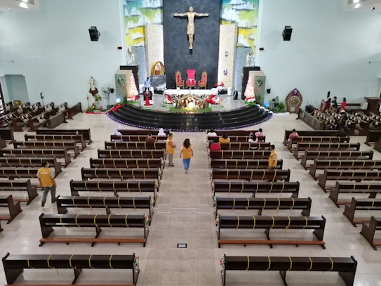 A large church interior featuring wooden benches with yellow ribbons, a central aisle, and a raised altar decorated with flowers and red cloth. A prominent statue of Jesus is on the wall behind the altar. A few people are seated in the pews while several others are standing and walking.