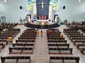 A large church interior featuring wooden benches with yellow ribbons, a central aisle, and a raised altar decorated with flowers and red cloth. A prominent statue of Jesus is on the wall behind the altar. A few people are seated in the pews while several others are standing and walking.