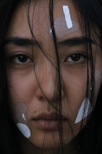 A close-up of a person's face featuring strands of dark hair partially obscuring their eyes and forehead. The skin has patches of white and beige makeup or paint applied in various shapes on the forehead and cheeks. The expression is neutral and introspective, and the lighting is dim, creating a moody atmosphere.